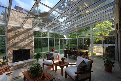 Interior of a sunroom with a dining room table, outdoor chairs and a coffee table, and a fireplace with a ceiling fan overhead. 